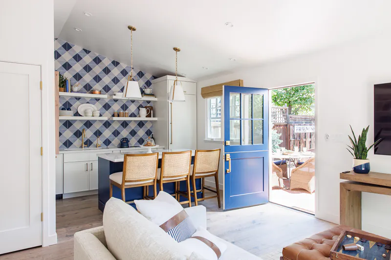 Small kitchen with white cabinets, a navy island with cane stools, a blue checked tile wall and a blue front door